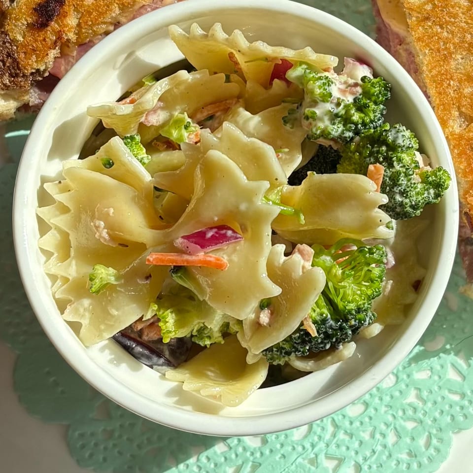 Close-up of a white bowl filled with creamy pasta salad, broccoli florets, and red onions.